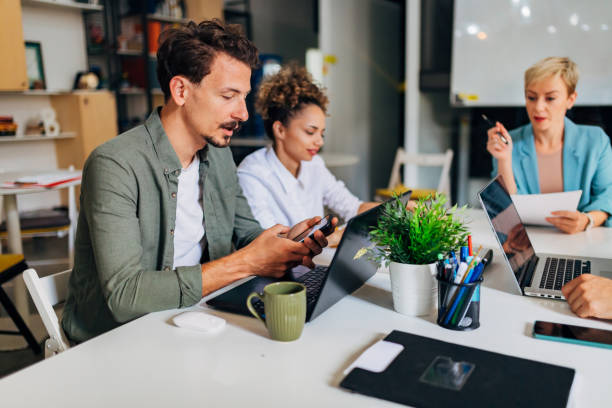 istock A man is sitting at a conference table with a group of colleagues. While learning how to code a program, he uses a laptop and a mobile phone 1432255671