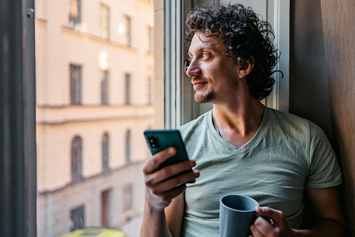 Handsome young man drinking coffee and using smart phone while leaning on a window sill.