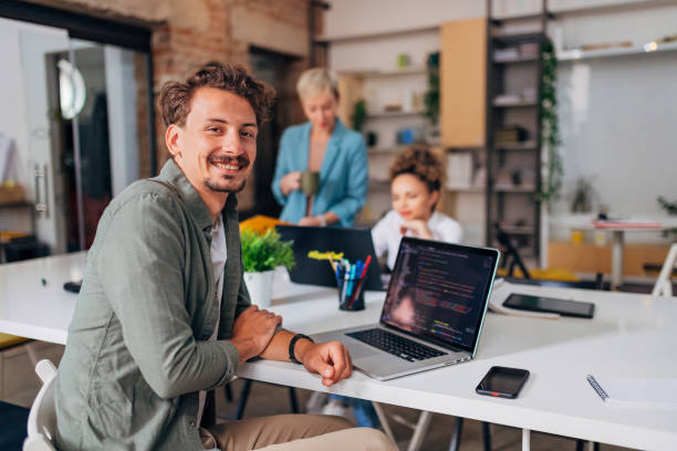 A cheerful man sits at a conference table across from his two female colleagues and learns to code Portrait of a smiling man learning coding techniques using a laptop and looking directly into the camera java stock pictures, royalty-free photos & images