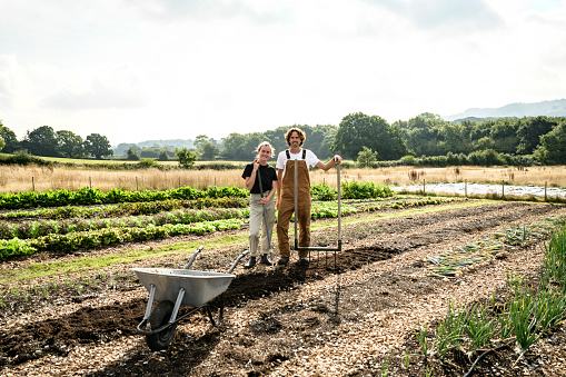 Full length view of man and woman in 20s and 30s pausing from preparing planting row with broadfork and compost to smile at camera.