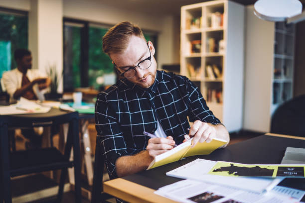 istock Concentrated male millennial making memo notes during learning knowledge project in coworking space for students, smart casual hipster guy writing test answer in paper textbook for education 1432273271