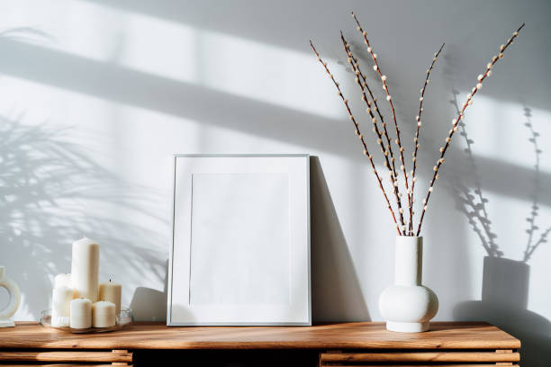 istock Modern minimalist style interior with white poster mockup, candles and blooming branches of the pussy willow in vase on wooden console under sunlight and shadows on a white gray wall. Selective focus 1432274104