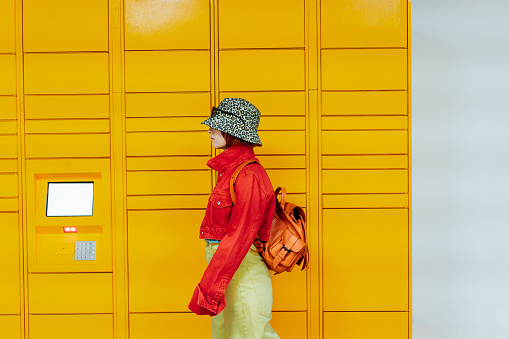 Hipster fashion young woman in bright clothes going past modern postal automatic mail terminal with self service device for receiving an order. Electronic locker for storing parcels. Selective focus