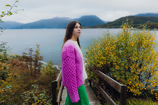 Happy woman with long hair and in a colorful sweater contemplating the fall season by walking at the pier by the sea with view of the beautiful mountain peaks in More og Romsdal county, Norway
