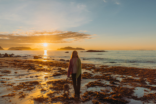 Happy female with long hair and in a colorful sweater admiring the bright  sunset at the beach with a cute pug, with view of the sea and the islands beyond the horizon in More og Romsdal, Norway