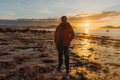 Happy male in a red jacket walking with a cute pug at the scenic beach with a view of the islands during bright sunset in More og Romsdal, Norway