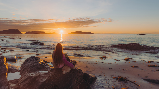 Happy female with long hair and in a colorful sweater admiring the bright sunset sitting with a pug at the beach with a view of the sea and the islands beyond the horizon in More og Romsdal, Norway