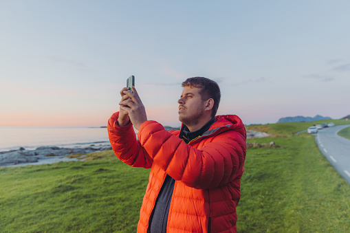 A man in red jacket taking photo of the scenic sunset at the seacoast of Norway
