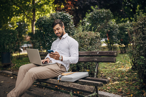 One man, modern businessman sitting on park bench, he is using laptop and smart phone.