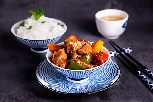 Chinese stir fried chicken with red, green, and yellow pepper bells served with rice. In the background of the photos there are jasmin tea, energy balls and prawn crackers. The food is served in elegant china and ceramic dishes.