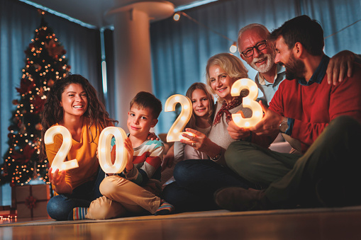 Happy family celebrating New Years Eve at home with kids, sitting by the Christmas tree, holding sparklers and illuminative numbers 2023 representing the upcoming New Year