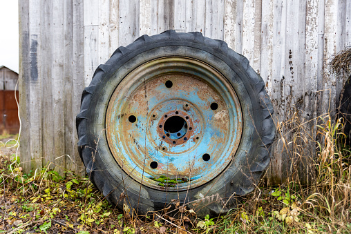 a large blue rusty wheel from a tractor stands near the wall of the barn among the grass