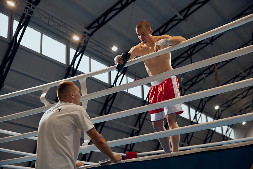 Strength, attack and motion concept. Shirtless muscular athlete in boxing gloves and shorts practicing punch at boxing ring at gym. Workout with personal coach, team trainer