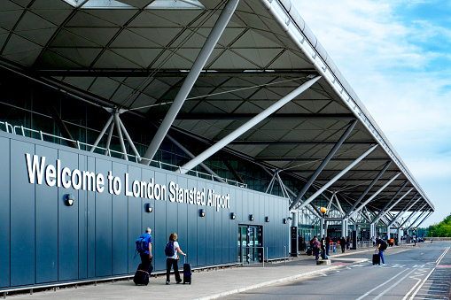 Passengers with luggage walking towards the entrance of London Stansted Airport with the welcome sign in the photo. Taken during the day in September 2022.