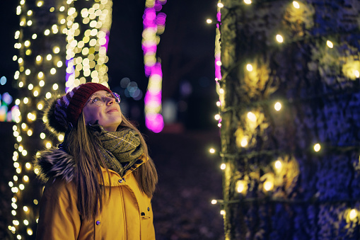 Teenage girl is looking at the Christmas lights in public park. The girl is standing by a tree and is looking up.\nCanon R5