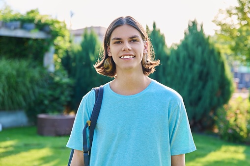 Outdoor portrait of smiling teenage guy with backpack looking at camera. Handsome young student male 18, 19 years old, with hairstyle, summer nature background