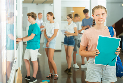 Smiling confident teenage girl student standing with backpack on college campus, holding folder with blank space for text, copyspace