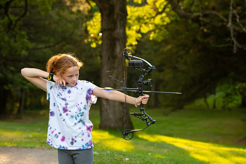 Side view of a young girl practicing at an outdoor archery range. She has just released the arrow, which can be seen flying through the air.