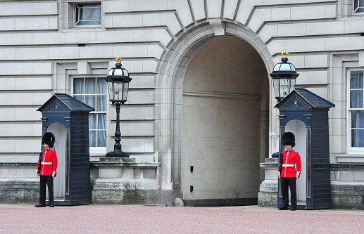 Armed soldiers guards using bearskin standing in the front yard at Buckingham Palace, London, England. This picture was taken from outside the palace during the changing of The Queen's Guard, the infantry soldiers charged with guarding the official royal residences in the United Kingdom. A bearskin is a tall fur cap, usually worn as part of a ceremonial military uniform.