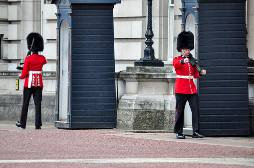 Armed soldiers guards using bearskin are marching in the front yard at Buckingham Palace, London, England. This picture was taken from outside the palace during the changing of The Queen's Guard, the infantry soldiers charged with guarding the official royal residences in the United Kingdom. A bearskin is a tall fur cap, usually worn as part of a ceremonial military uniform.