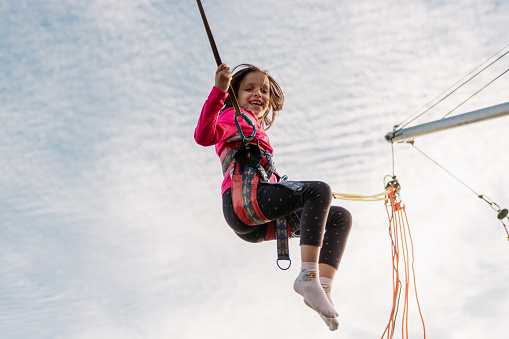 Girl is jumping on a bungee trampoline. A child with insurance and stretchable rubber bands hangs against the sky. The concept of happy childhood and games in the amusement park.