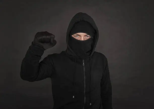 Man in the black hoody with hood wearing balaclava mask with fist up on dark background