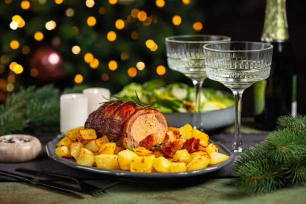 Christmas or New Year dinner table. Close-up of baked duck roulade stuffed with meat and oranges, baked potato, salad, champagne coupe glasses in front of Christmas tree and burning candles.