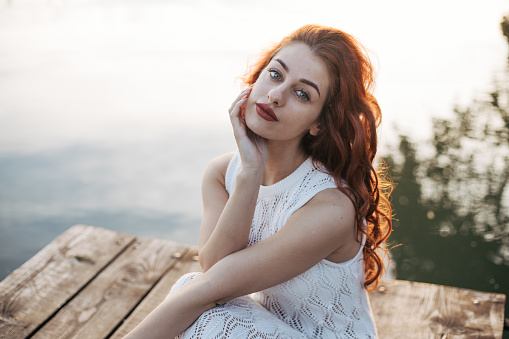 Young woman with red curly hair is posing in white lace dress on the float with sunlight in the backgorund. Barefoot in beautiful nature with little makeup makes great photoshoot like in a fairytale.