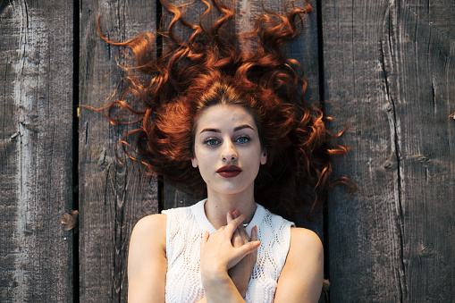 Young woman with red curly hair is posing in white lace dress on the float with sunlight in the backgorund. Barefoot in beautiful nature with little makeup makes great photoshoot like in a fairytale.