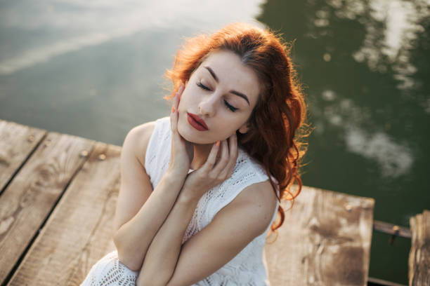 Young woman with red curly hair is posing in white lace dress on the float with sunlight in the backgorund. Barefoot in beautiful nature with little makeup makes great photoshoot like in a fairytale.