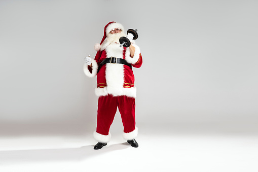 Full length photo of real Santa Claus doing fit exercise. Christmas time. Xmas vibes. A lot of copy space on the light gray studio background.