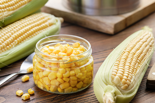 Pickled sweet corn in a glass jar, fresh corncobs, saucepan on kitchen table.