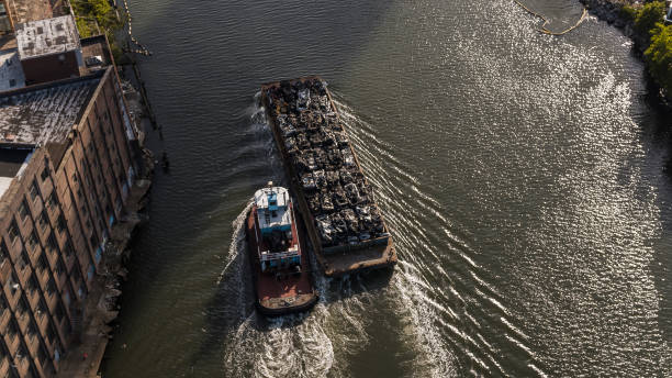 A tugboat pushes a barge along the calm waters of the Elbe (Labe) River on an overcast day. The boat, with a red and white hull and a small cabin, is centered in the frame, creating a sense of motion as it heads towards the right. The river reflects the gray sky, adding a somber yet serene tone to the image. Bare trees line the riverbanks, hinting at a winter or early spring setting. In the background, a hill rises with patches of snow, and a smokestack and power lines are visible, suggesting an industrial presence in the landscape. This image captures the working river environment, blending natural and man-made elements with a muted, atmospheric palette. Ideal for themes related to transportation, industry, and river landscapes.