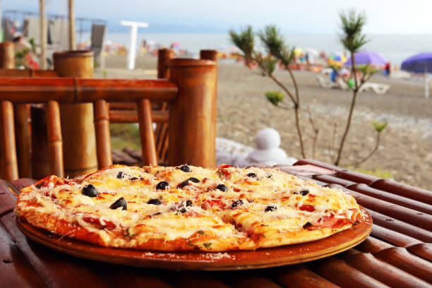 istock Big delicious pizza on a bamboo table. Pie tomato zucchini mozzarella, pesto and virgin olive oil. Angled view of the sea beach 1432396536