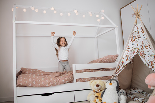 Cute little happy caucasian girl jumping on white scandinavian wooden house frame bed at home. Happy childs moments.