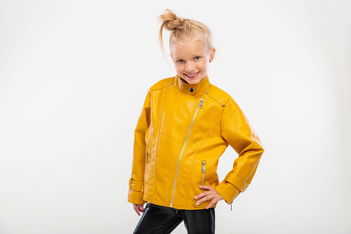 Pretty blonde little girl in a leather mustard jacket, black snake texture leggings and boots. High fashion, standing pose, isolated against a studio background