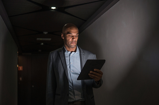 Portrait of serious mixed race business man in early 40s standing in a dark hotel corridor and using a digital tablet.