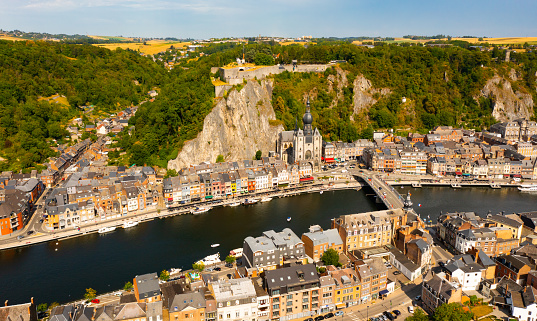 Aerial view of Walloon city of Dinant located along Meuse river with Gothic medieval Collegiate Church of Our Lady and fortified castle perched atop green vertical cliff in summer,  Belgium