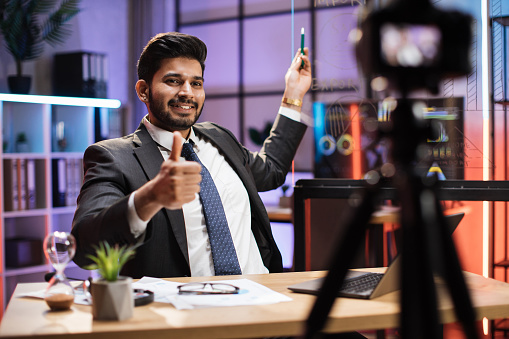 Concept of distant work at night. Attractive confident arab skilled businessman explaining online economic charts on glass board to his colleagues sitting in front of video camera showing thumb up.