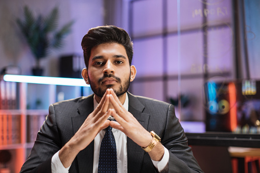 Indian bearded man in formal suit sitting at desk with modern laptop and looking at camera with serious facial expression. Concept of people and office work.