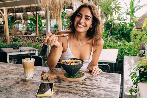 Charming smiling female in white top enjoying delicious breakfast in outdoor cafeteria and looking at camera while spending vacation in exotic country