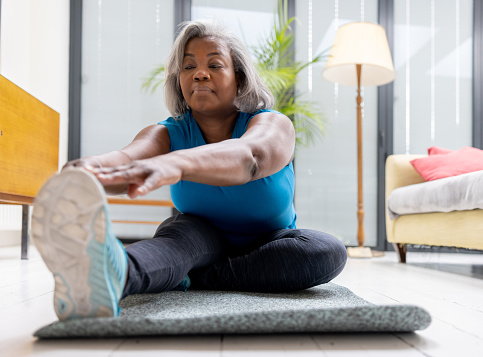 Mature black woman exercising at home and stretching while sitting on a mat - fitness concepts