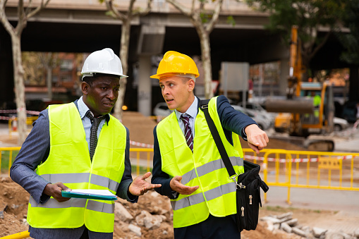 Two engineers, Caucasian and African-american men in hardhats and warnvests, standing and talking about road paving works. One man pointing with finger on something.
