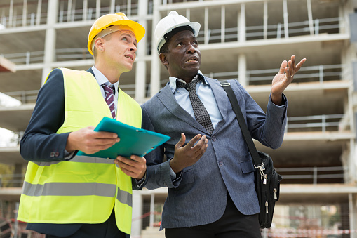 European foreman and African-american engineer in hardhats standing on building site and talking about construction.