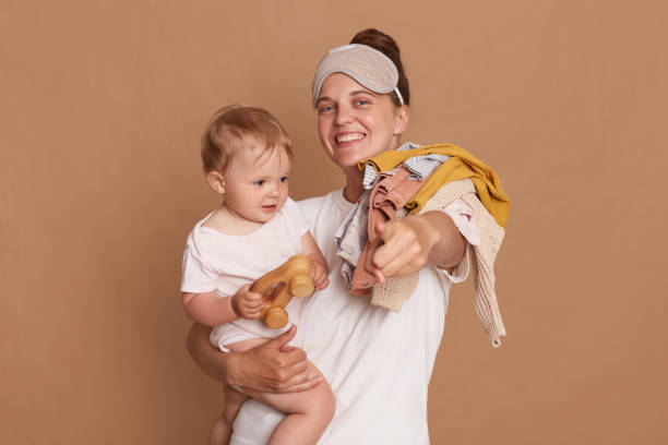 portrait d’une femme heureuse et positive portant un t-shirt blanc et un masque de sommeil, mère avec sa petite fille tenant du linge sur l’épaule, pointant du doigt la caméra, isolée sur fond brun. - caméra à lépaule photos et images de collection