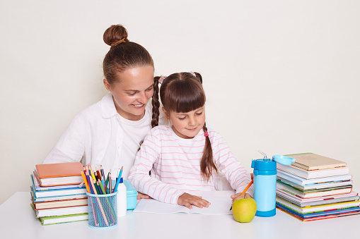 Indoor shot of attractive mother with her daughter sitting at table with books doing homework, mommy or tutor helping to pretty cute schoolkid to tasks, posing isolated over white background.