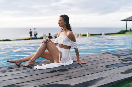 Full length of peaceful slim barefoot female in white swimwear looking away while resting on poolside against cloudy sky on resort