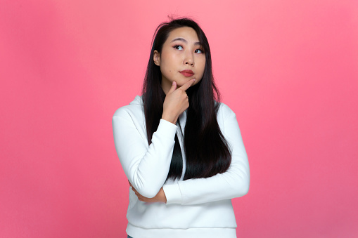 Pensive puzzled young asian girl looking up, considering problem solution, pondering decision on pink studio background. High quality photo