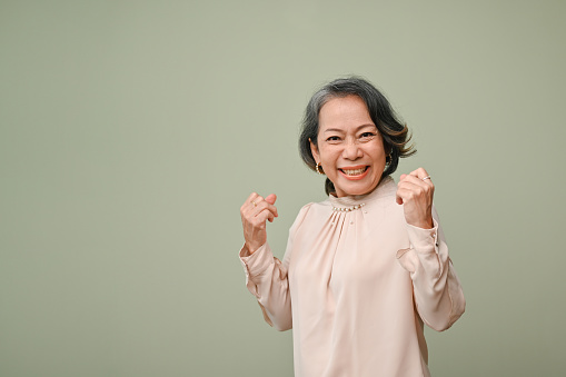 Joyful 60s aged-asian woman raises hands with clenched fists, standing against green background, celebrating her success, winning, goal achievement, victory, excitement,  good energy, say yes.