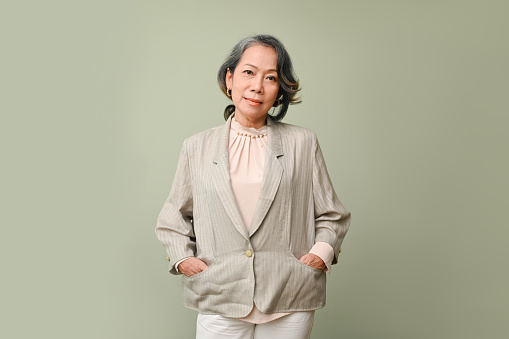 Professional and successful aged-asian businesswoman or female CEO in casual suit hands in suit pockets, standing isolated over green studio background.